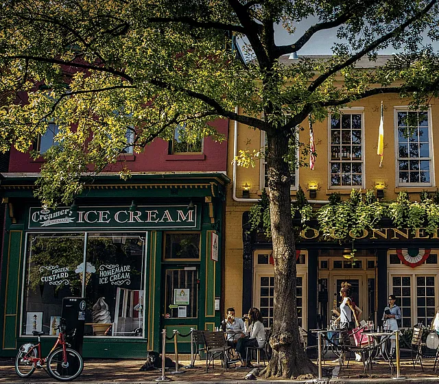 Charming urban streetscape featuring a vibrant ice cream shop and a bustling café with outdoor seating beneath lush greenery.