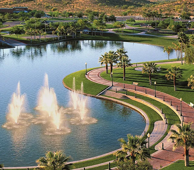Serene lake view with elegant fountains, surrounded by lush greenery, palm trees, and scenic pathways.