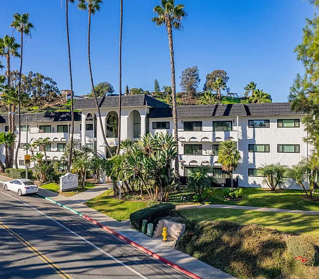 Charming apartment building surrounded by lush palm trees and vibrant greenery, offering a serene urban oasis with easy street accessibility.