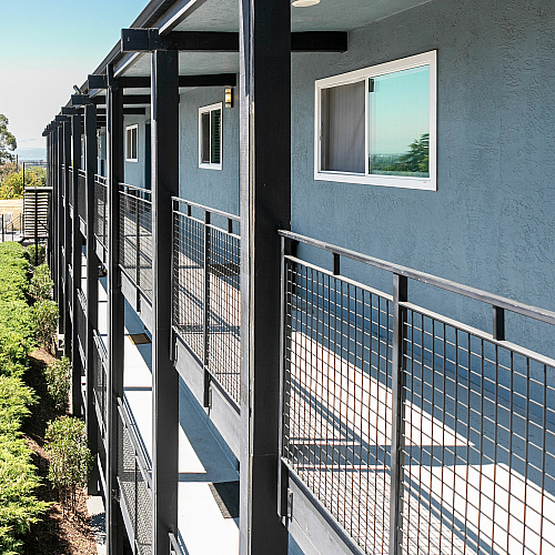 Modern apartment exterior with sleek black railings and stylish blue facade, featuring a sunlit walkway and lush greenery.