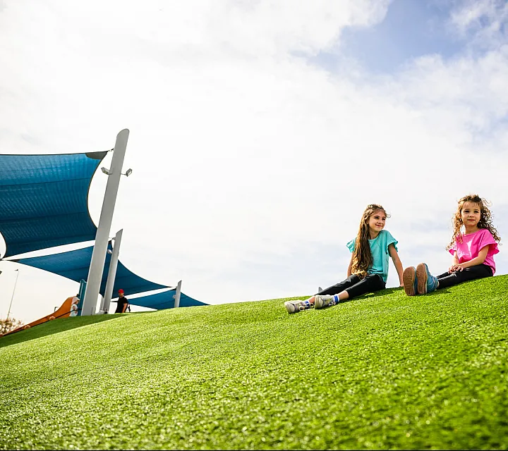 Sunny park scene with two children in colorful outfits sitting on vibrant green grass under a clear blue sky.