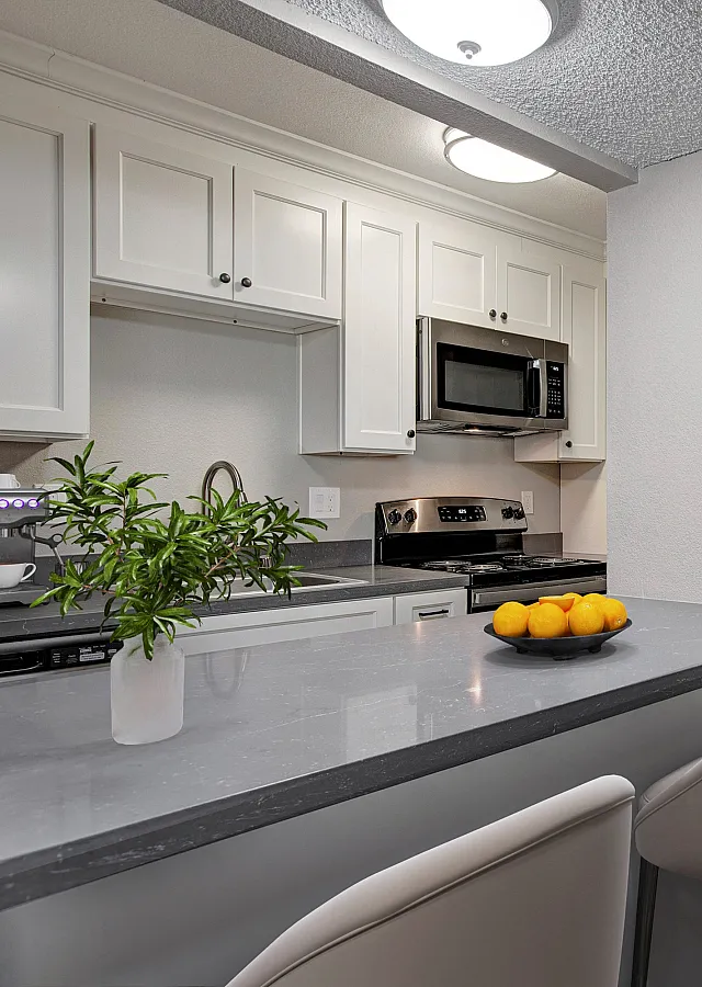 Modern kitchen with sleek gray countertops, elegant white cabinetry, and a cozy breakfast bar. Hallway leads to a warmly lit bedroom, enhancing the inviting atmosphere.