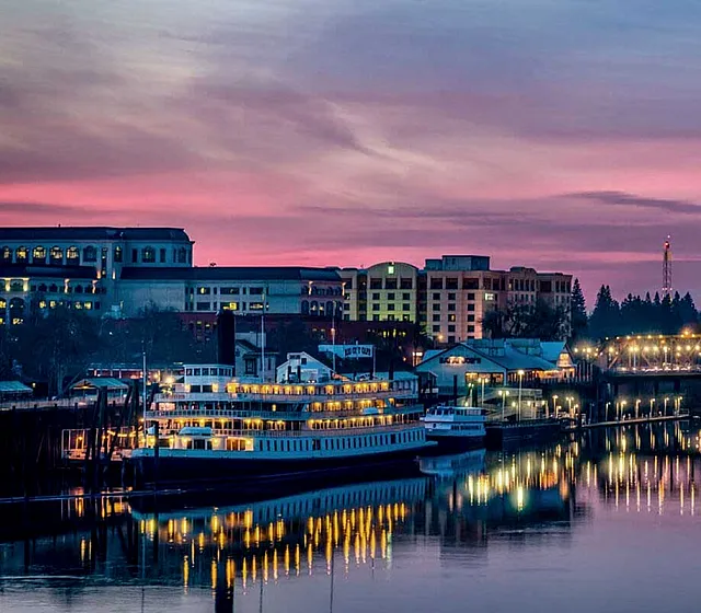 Captivating cityscape view featuring a beautifully illuminated tower and riverfront buildings at sunset, reflecting vibrant colors over tranquil waters.
