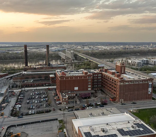 Scenic aerial view of industrial architecture with a vibrant sunset, highlighting a blend of historic brick buildings and lush riverside landscape.