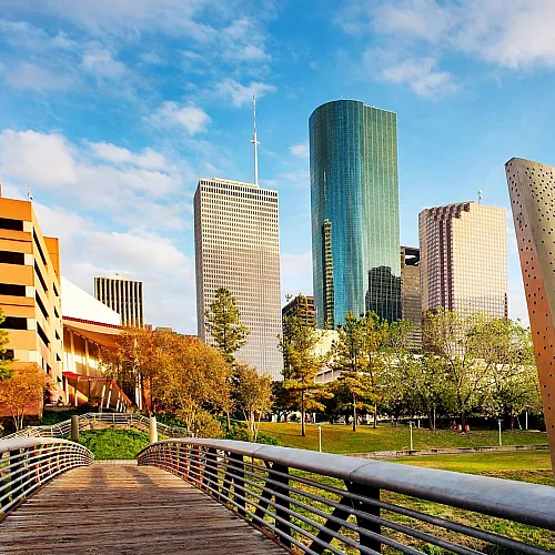 Vibrant cityscape with modern skyscrapers and lush green park, connected by a scenic pedestrian bridge under a clear blue sky.