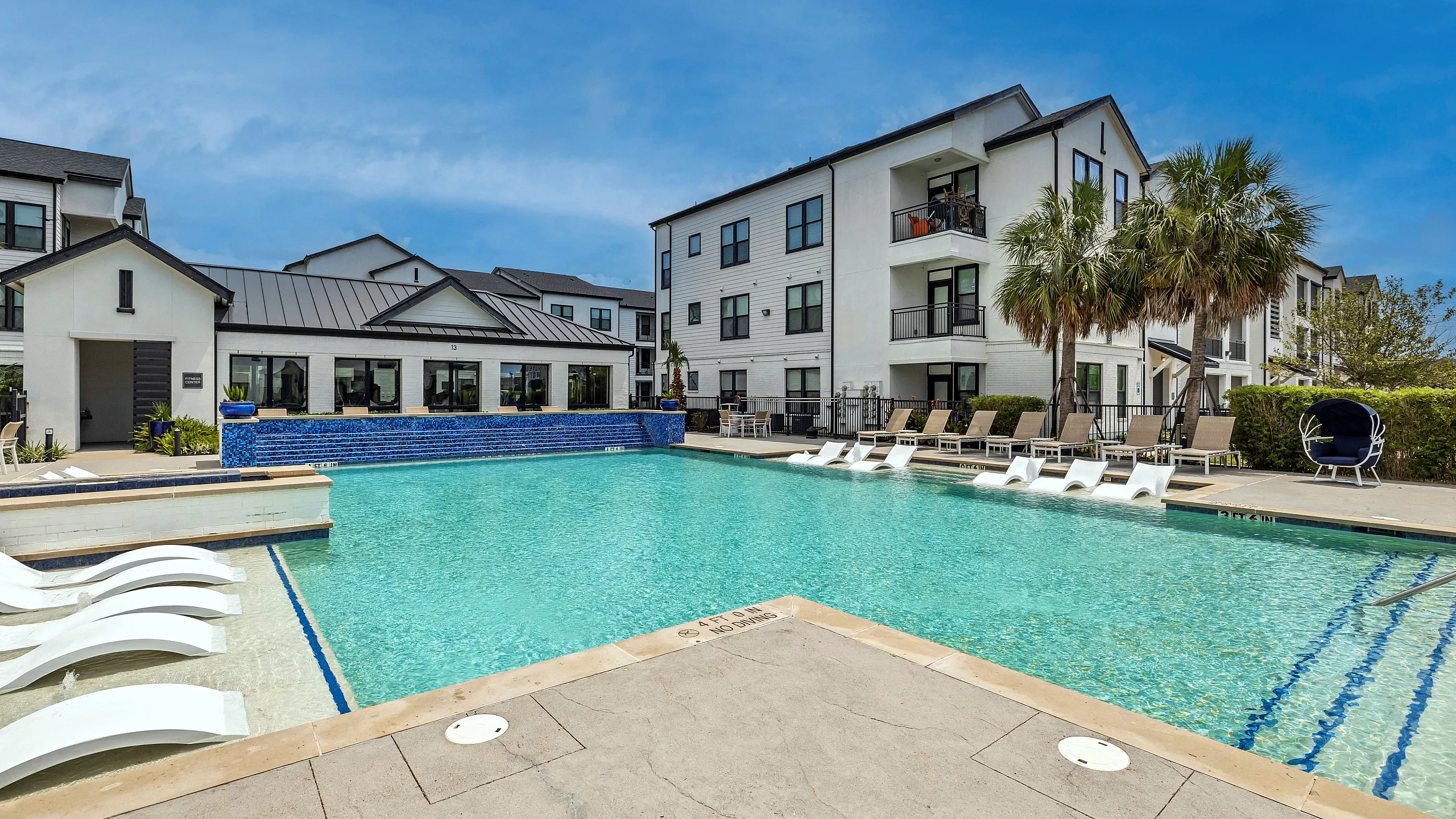 Luxury apartment complex with a sparkling pool, modern sun loungers, and stylish building exteriors surrounded by palm trees.