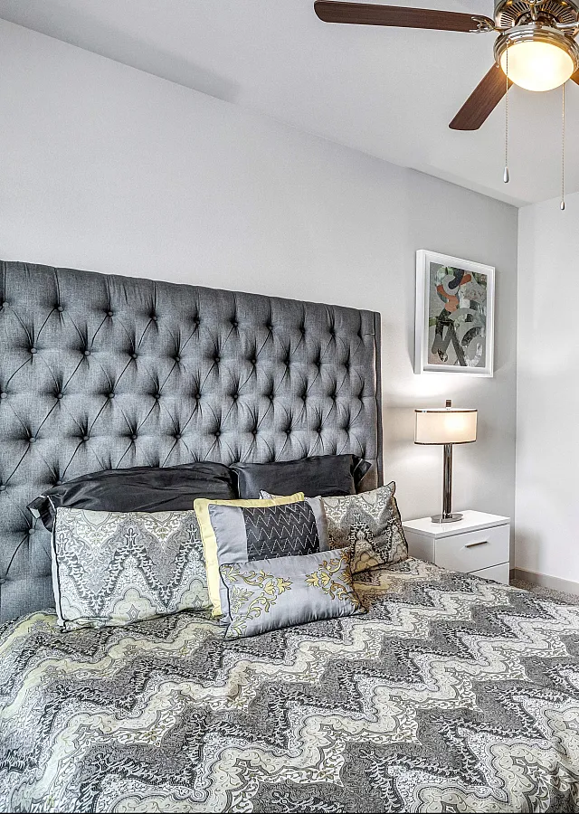 Chic bedroom with plush gray headboard, elegant patterned bedding, and a modern ceiling fan, adjacent to a stylish bathroom in a cozy apartment setting.