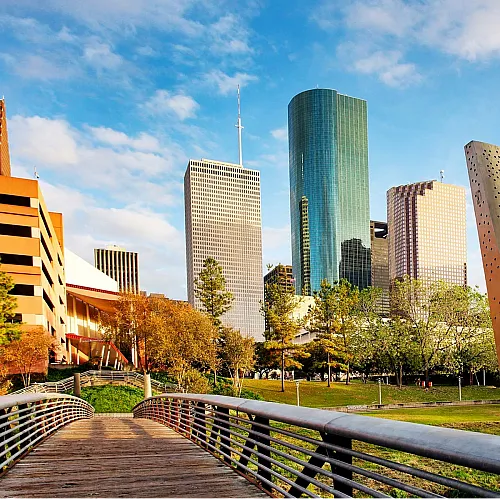 Vibrant cityscape with modern skyscrapers under a clear blue sky, featuring a scenic pedestrian bridge and lush greenery.