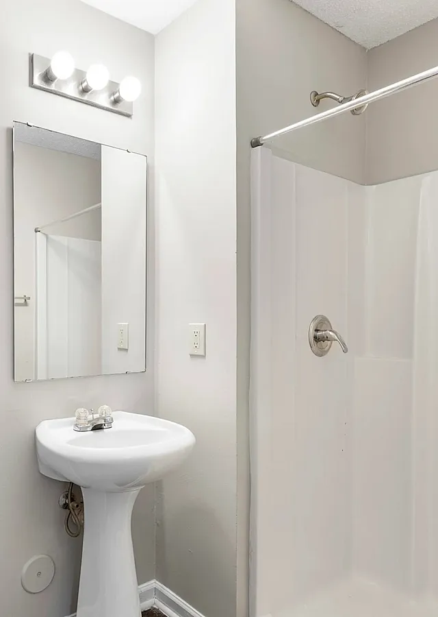 Bright and modern bathroom with a sleek pedestal sink, elegant mirror, wooden cabinet storage, and a pristine walk-in shower.