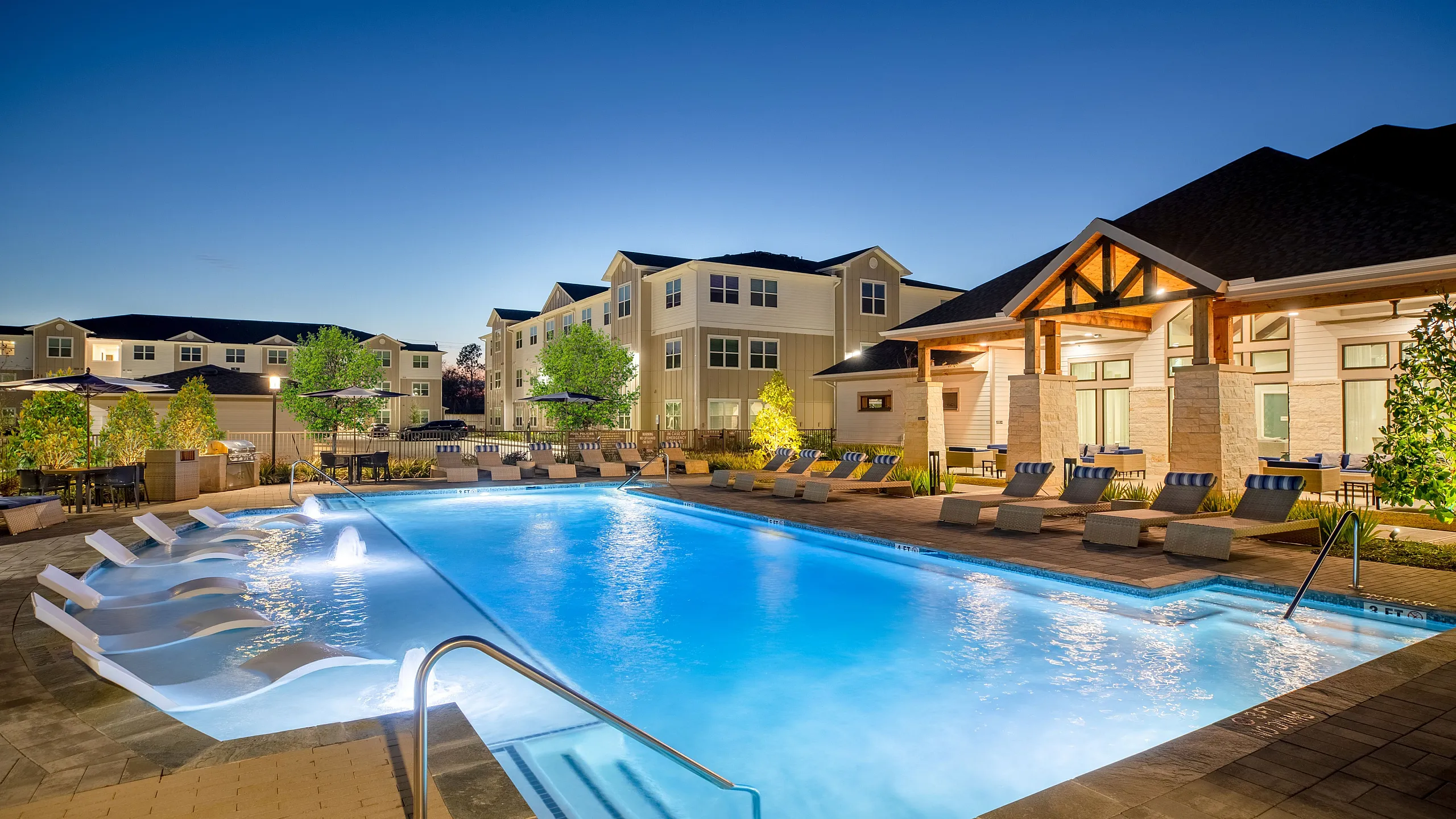 Luxurious apartment complex showcasing a sparkling swimming pool, surrounded by elegant deck furniture and modern architecture, illuminated under the evening sky.