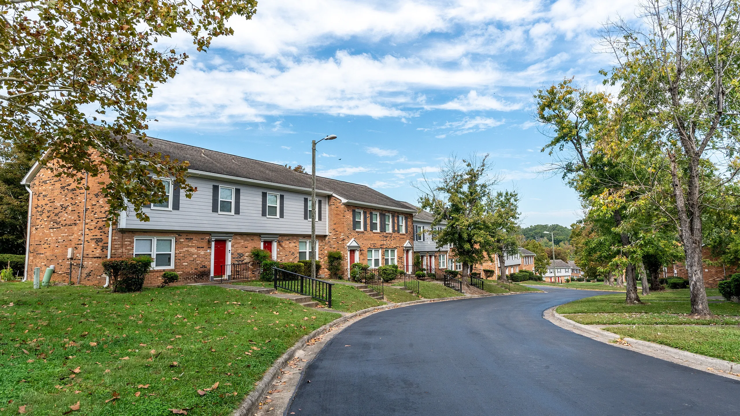 Charming suburban neighborhood with brick townhouses, lush green lawns, and a tree-lined street under a bright blue sky.