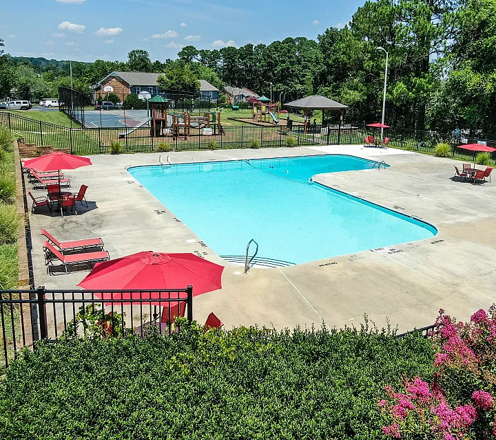 Inviting community pool area with vibrant red umbrellas, lounge chairs, and a backdrop of lush greenery.