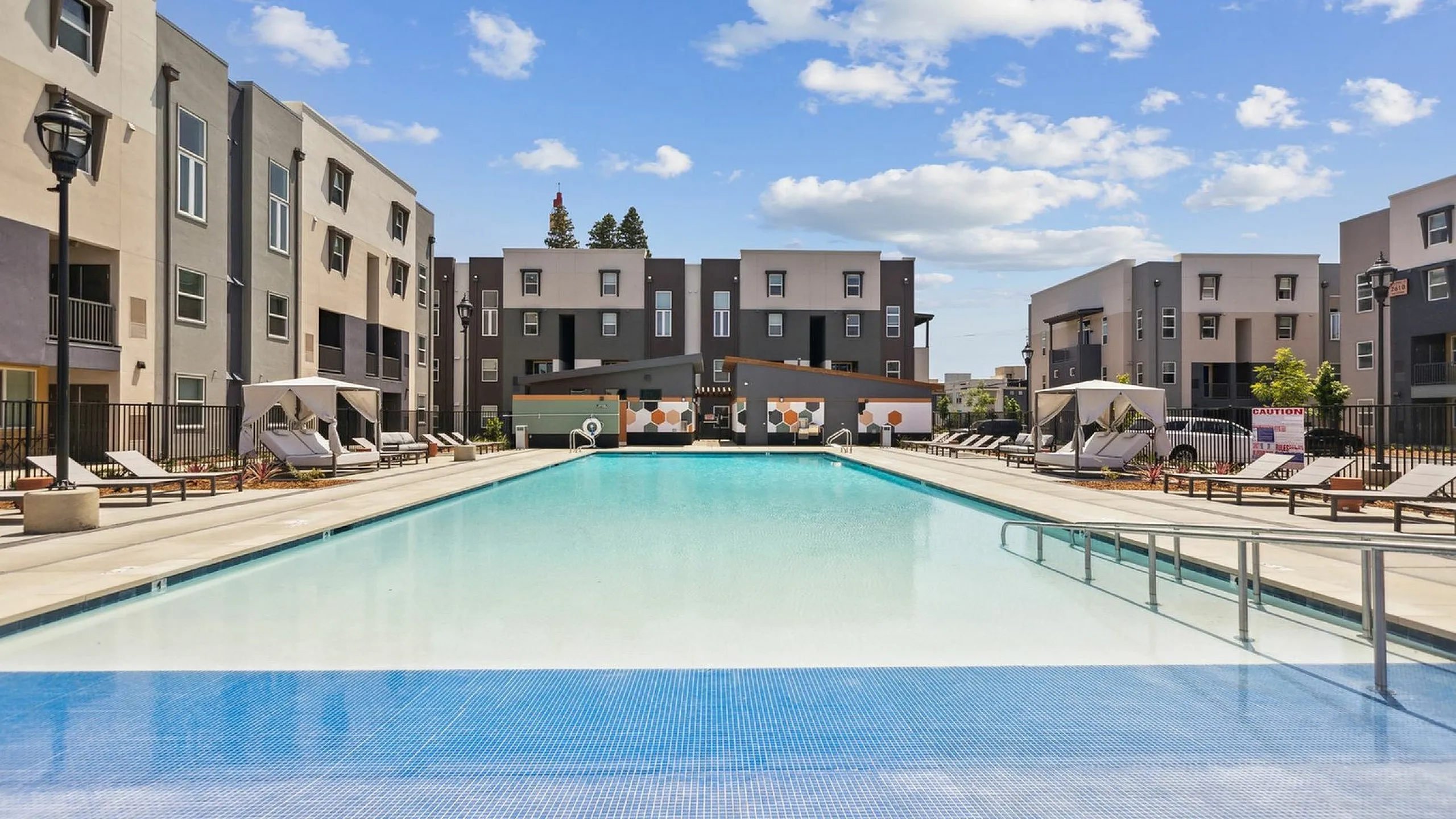 Modern apartment complex featuring a sparkling swimming pool, sun loungers, and stylish architecture under a clear blue sky.