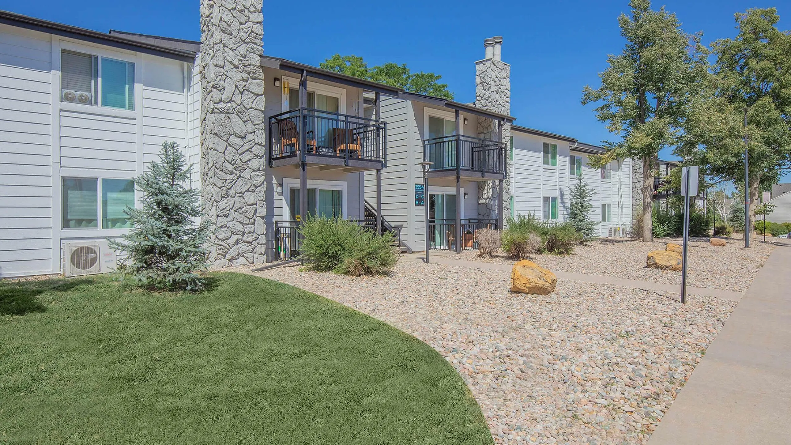 Charming apartment complex with modern stone accents, lush green lawn, and cozy private balconies under a clear blue sky.