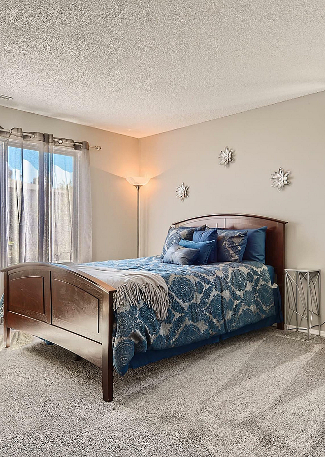 Bright and inviting bedroom with elegant d&eacute;cor, featuring a cozy bed with a stylish blue bedspread, large windows with flowing curtains, and a plush blue armchair enhancing the luxurious ambiance.