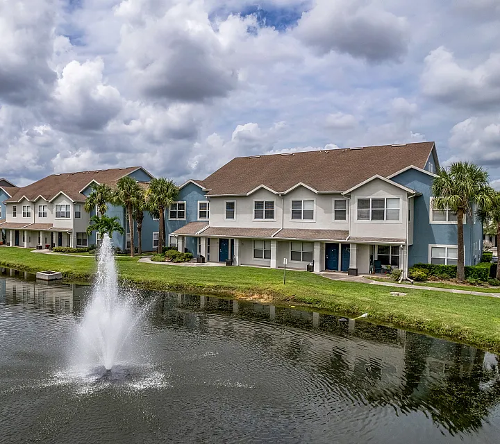 Charming waterfront townhomes with palm-lined paths and a serene fountain under beautiful clouded skies.