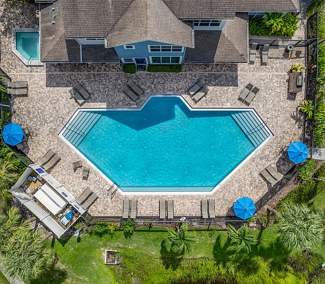 Aerial view of a luxurious hexagonal swimming pool surrounded by a spacious sun deck with lounge chairs, blue umbrellas, and lush green landscaping.