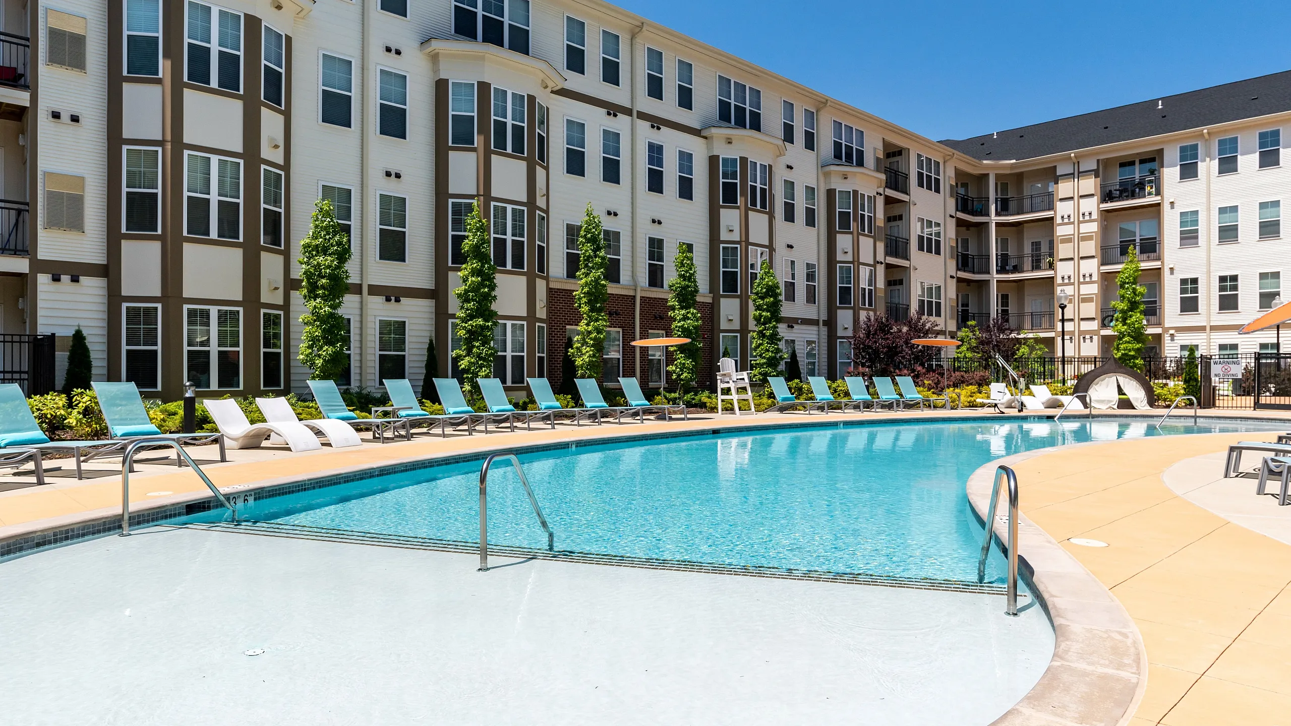 Luxurious apartment complex featuring a stunning outdoor pool with stylish lounge chairs, surrounded by beautifully landscaped greenery.