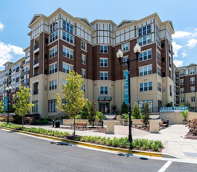Modern apartment building with a charming brick facade and large windows, set on a lush landscaped street, inviting with a 'Lease Today' sign.