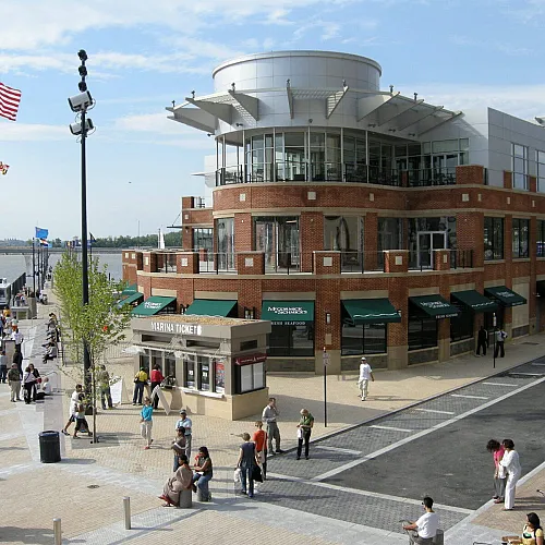 Vibrant waterfront plaza with brick building, marina ticket booth, and lively people enjoying the scenic riverside setting.
