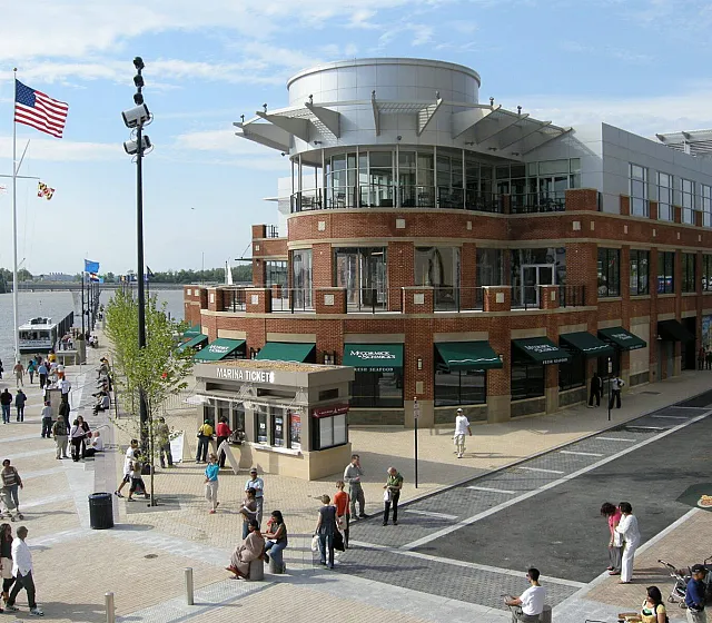 Vibrant waterfront scene with bustling people, modern brick building with green awnings, and waving American flags, perfect for dynamic urban living.