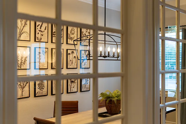 Elegant dining room with artistic botanical prints and a chic modern chandelier, viewed through French doors.