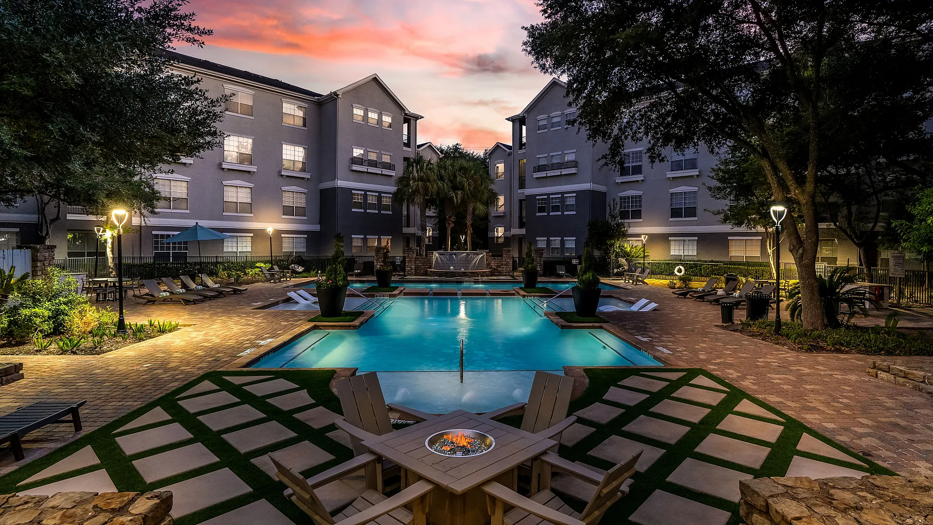 Luxurious apartment complex with a beautifully lit pool area and cozy outdoor seating, surrounded by lush greenery and charming multi-story buildings against a vibrant sunset sky.