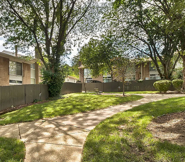 Charming apartment community with brick buildings, lush green landscaping, and winding pathways under mature trees.