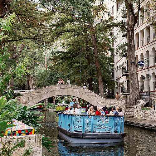 Charming riverside scene with a lively tour boat gliding under a picturesque stone bridge surrounded by lush greenery and historic architecture.