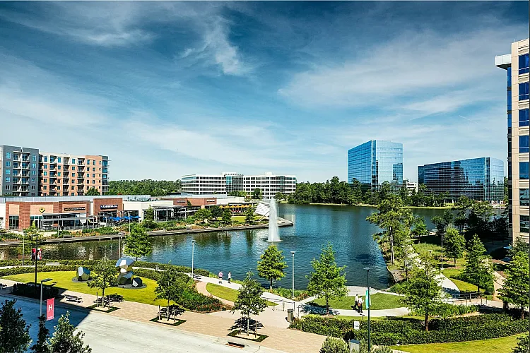 Vibrant cityscape featuring modern office buildings, a serene lake with a fountain, and lush green parks under a clear blue sky.