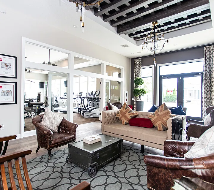 Modern apartment lounge with stylish seating, chic chandelier, and natural light from large windows, adjacent to a fitness room.
