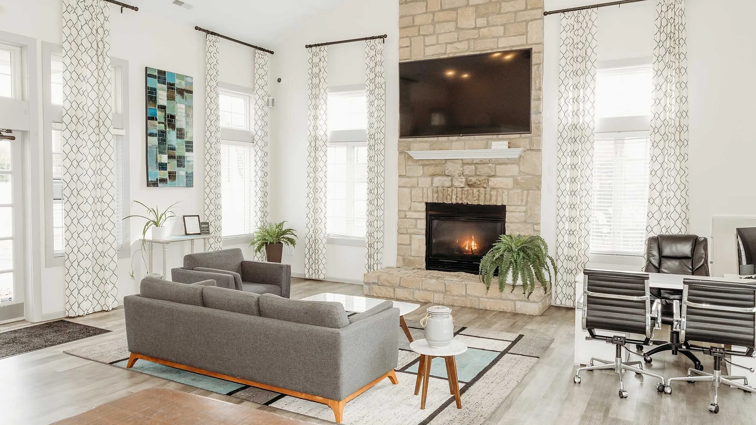 Inviting and modern living room featuring a cozy stone fireplace, stylish gray sofas, and sleek office chairs, all enhanced by abundant natural light and tasteful decor.