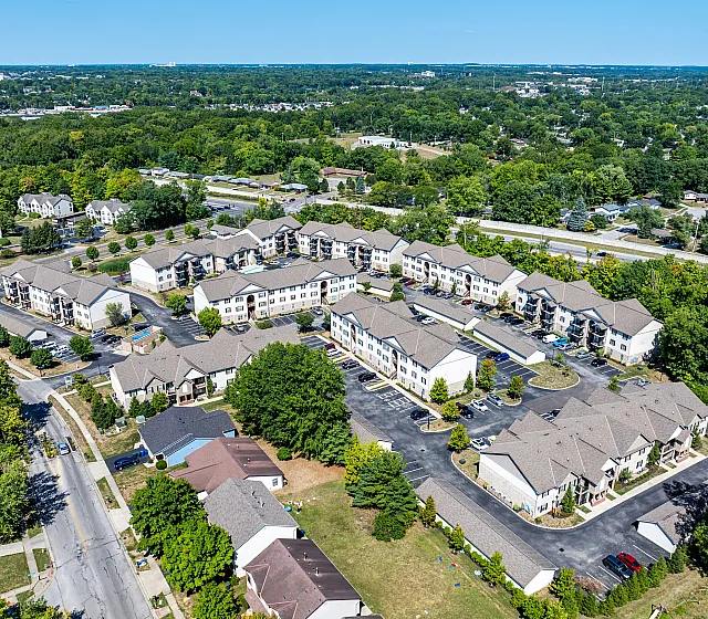 Expansive aerial view of a well-designed apartment complex nestled in lush greenery, showcasing multiple buildings with ample parking and tree-lined streets.