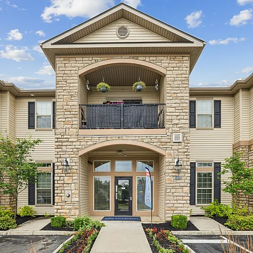 Charming apartment exterior with stone facade and inviting balcony, surrounded by lush landscaping under a bright blue sky.