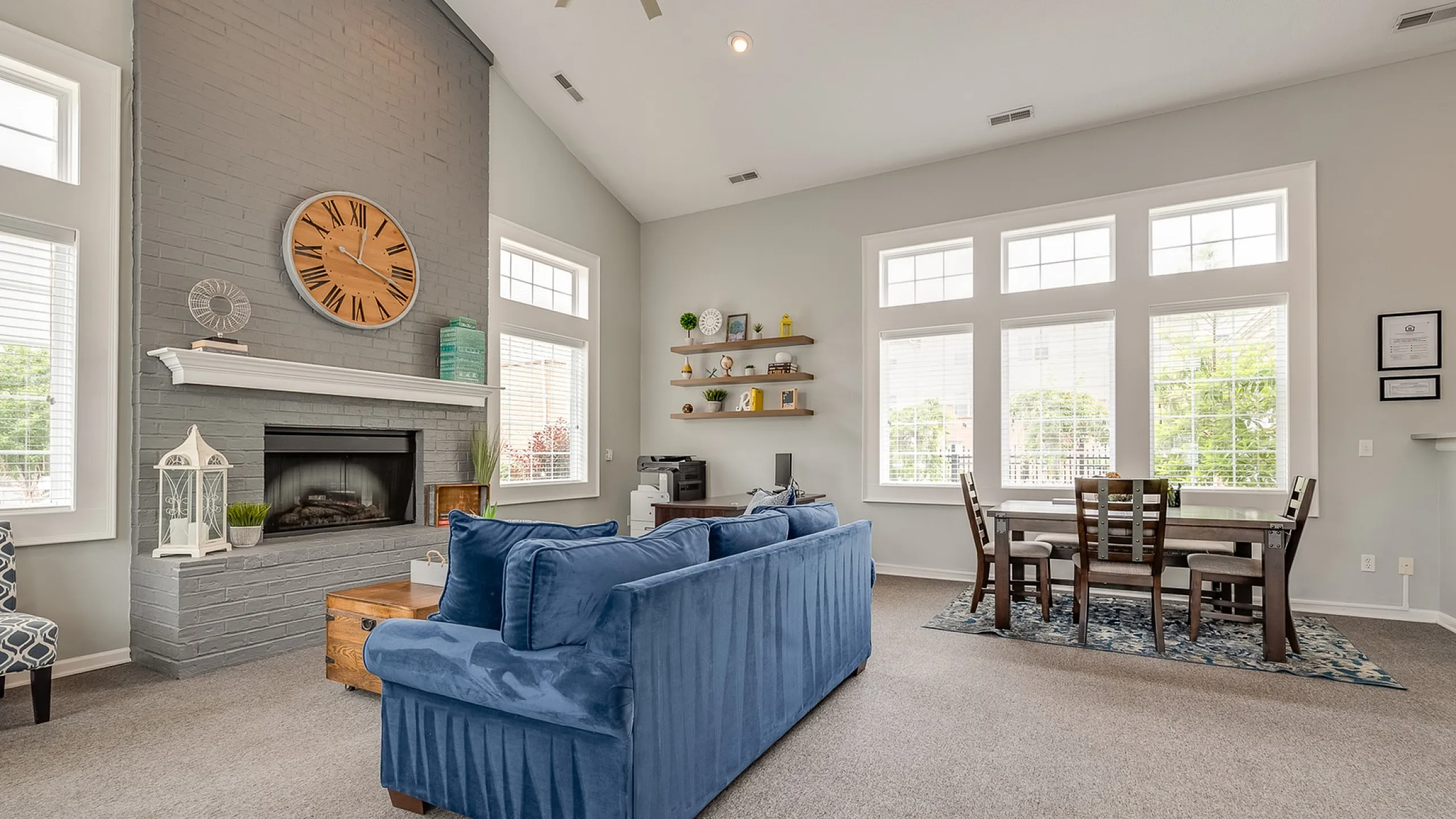 Inviting living room with a cozy blue sofa, elegant fireplace, and dining area beneath large, bright windows.