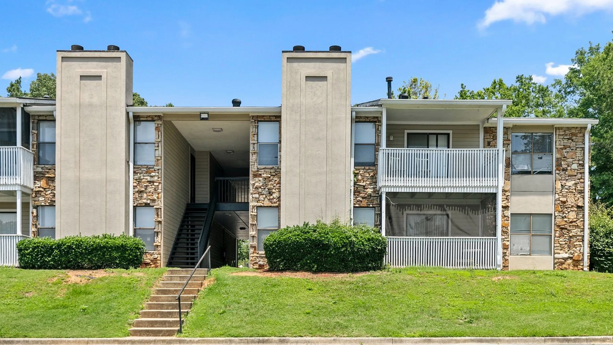 Inviting apartment building with charming stone facade and spacious balconies, surrounded by lush greenery and blue skies.