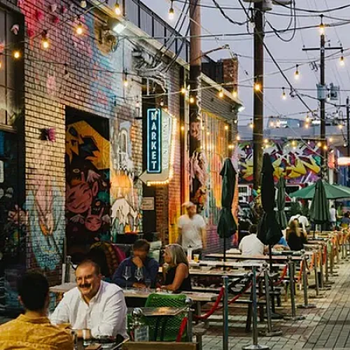 Vibrant urban alley with colorful murals, hanging lights, and lively outdoor dining at a bustling market setting.