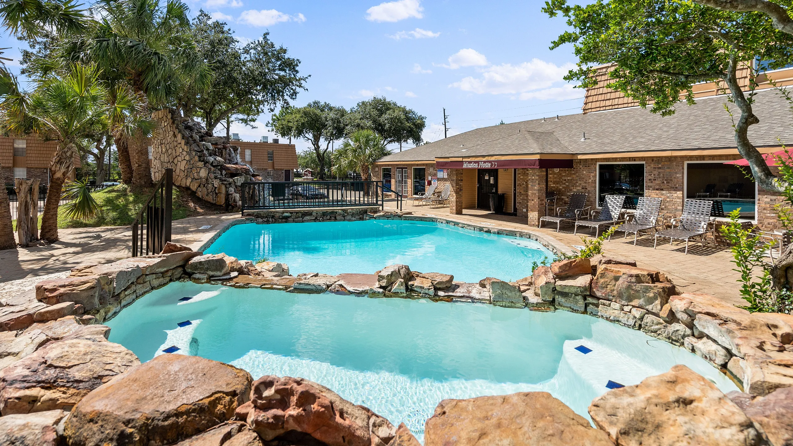 Inviting outdoor swimming pool with natural stone features, surrounded by lush greenery and lounge chairs under a sunny blue sky.