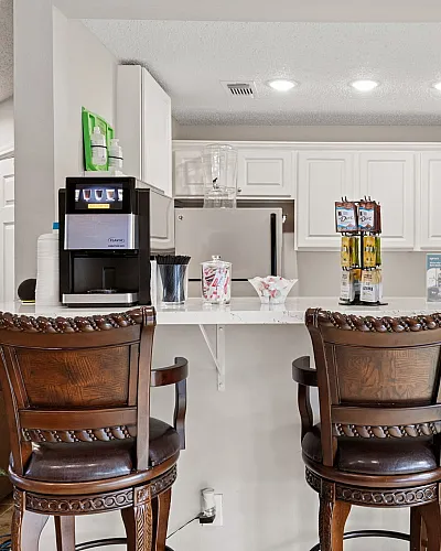 Inviting kitchen space featuring elegant wooden bar stools, a sleek coffee dispenser, and bright white cabinetry, accented with tasteful wall art.