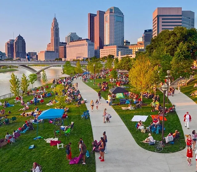 Vibrant riverfront park scene with people enjoying leisure activities on a sunny day. Lush greenery, modern cityscape, and scenic bridges in the background.