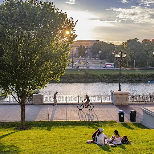 Serene riverside park with people enjoying a sunny day, featuring lush greenery, a cyclist, and an urban backdrop with flags and a colorful play area.