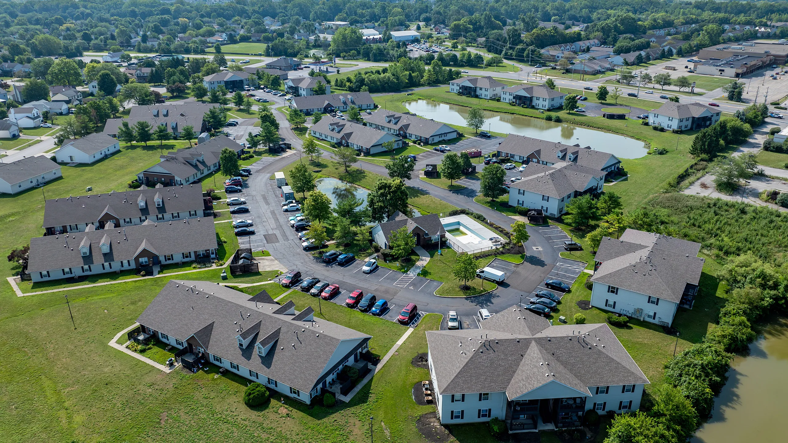 Scenic aerial view of a charming apartment community surrounded by lush greenery, featuring spacious units, ample parking, and a serene pond.