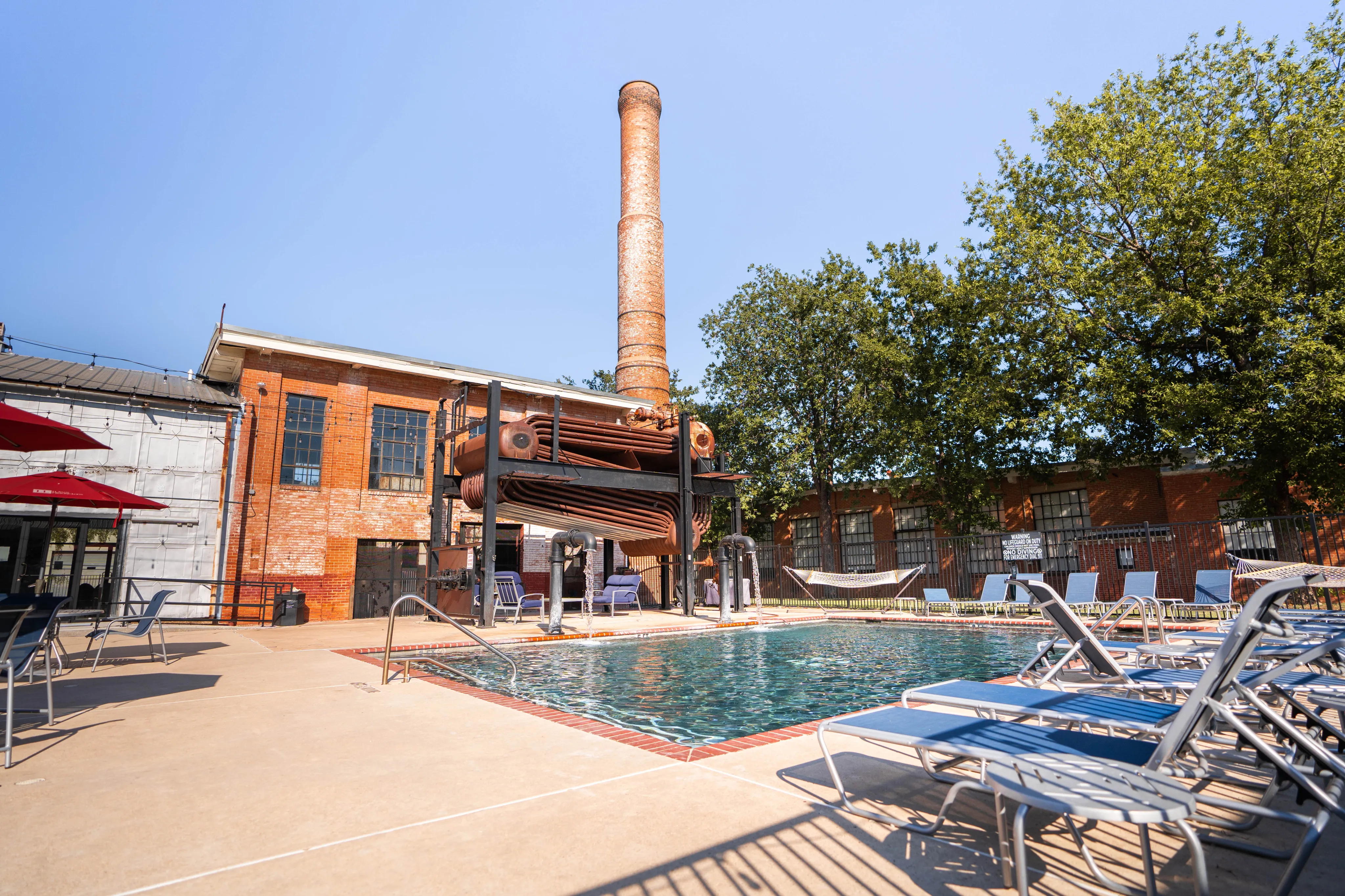 Inviting outdoor pool area with industrial charm, featuring lounge chairs, red brick building, and tall smokestack.