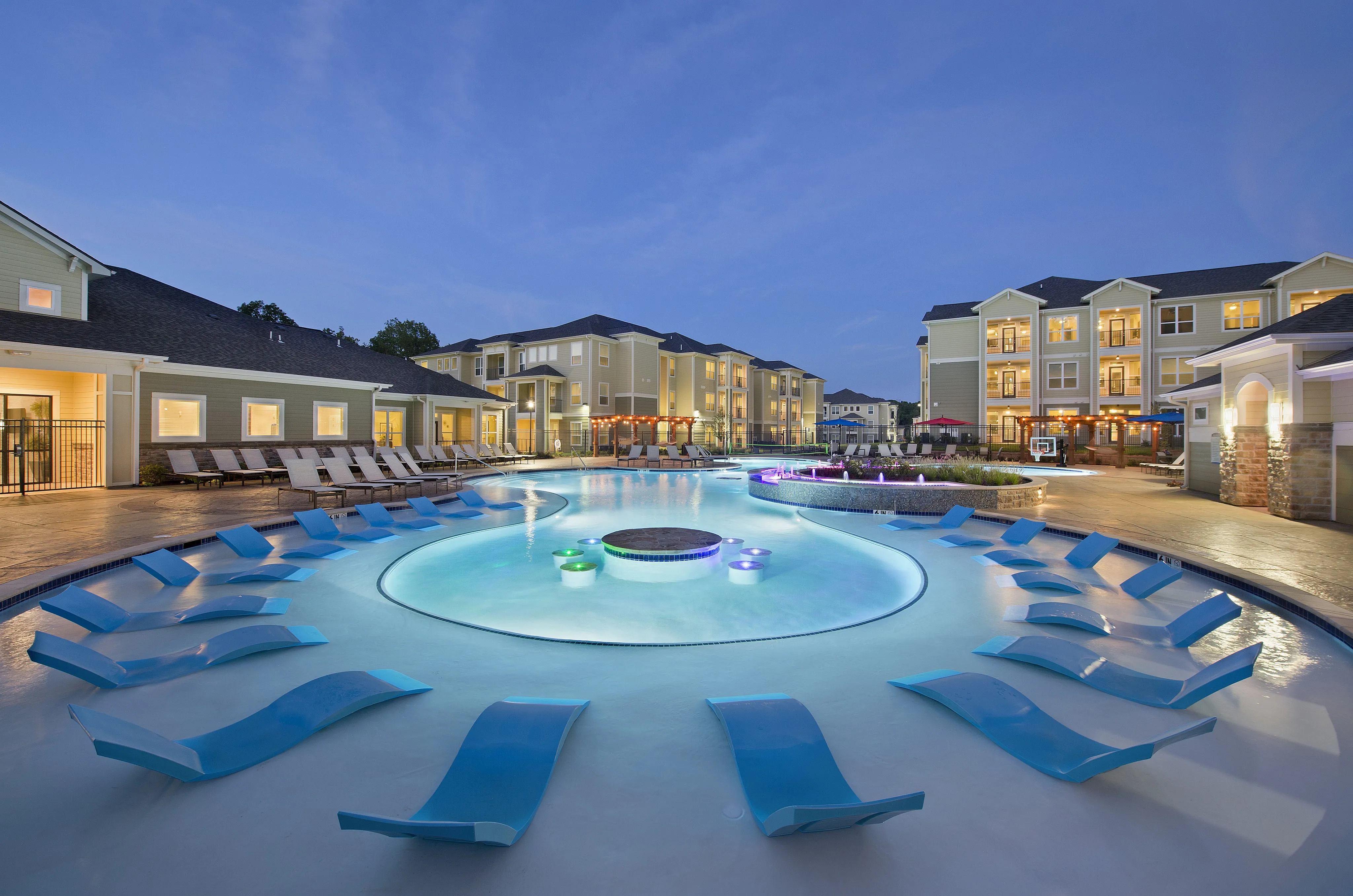 Luxurious apartment complex with a stunning resort-style pool surrounded by modern, spacious buildings under a beautiful twilight sky.