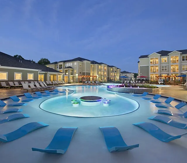 Luxurious resort-style apartment complex showcasing an elegant swimming pool with modern lounge chairs, surrounded by beautifully lit contemporary buildings at dusk.