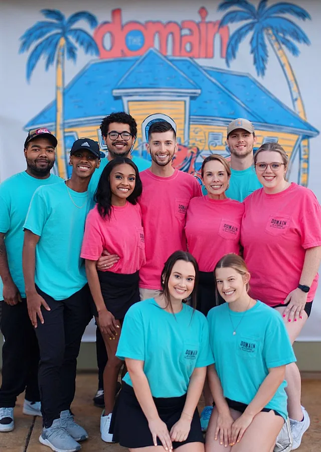 Cheerful group of people in branded T-shirts standing together in front of a vibrant and inviting house illustration, showcasing a lively community atmosphere.