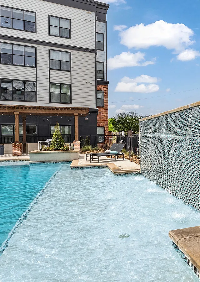 Inviting apartment pool area with a modern waterfall feature, surrounded by vibrant outdoor seating and lush greenery under a clear blue sky.