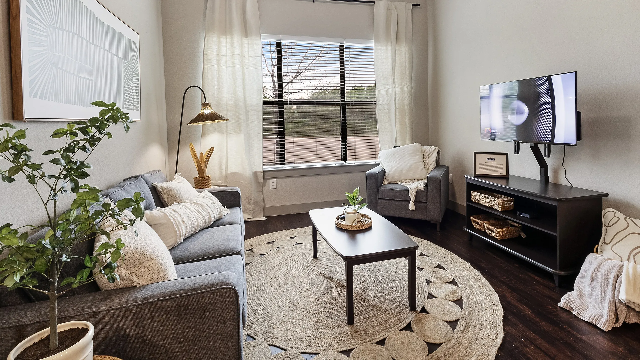Charming living room with elegant decor, featuring a cozy grey couch, stylish armchair, and modern TV setup. Lush greenery and natural light enhance the welcoming ambiance.