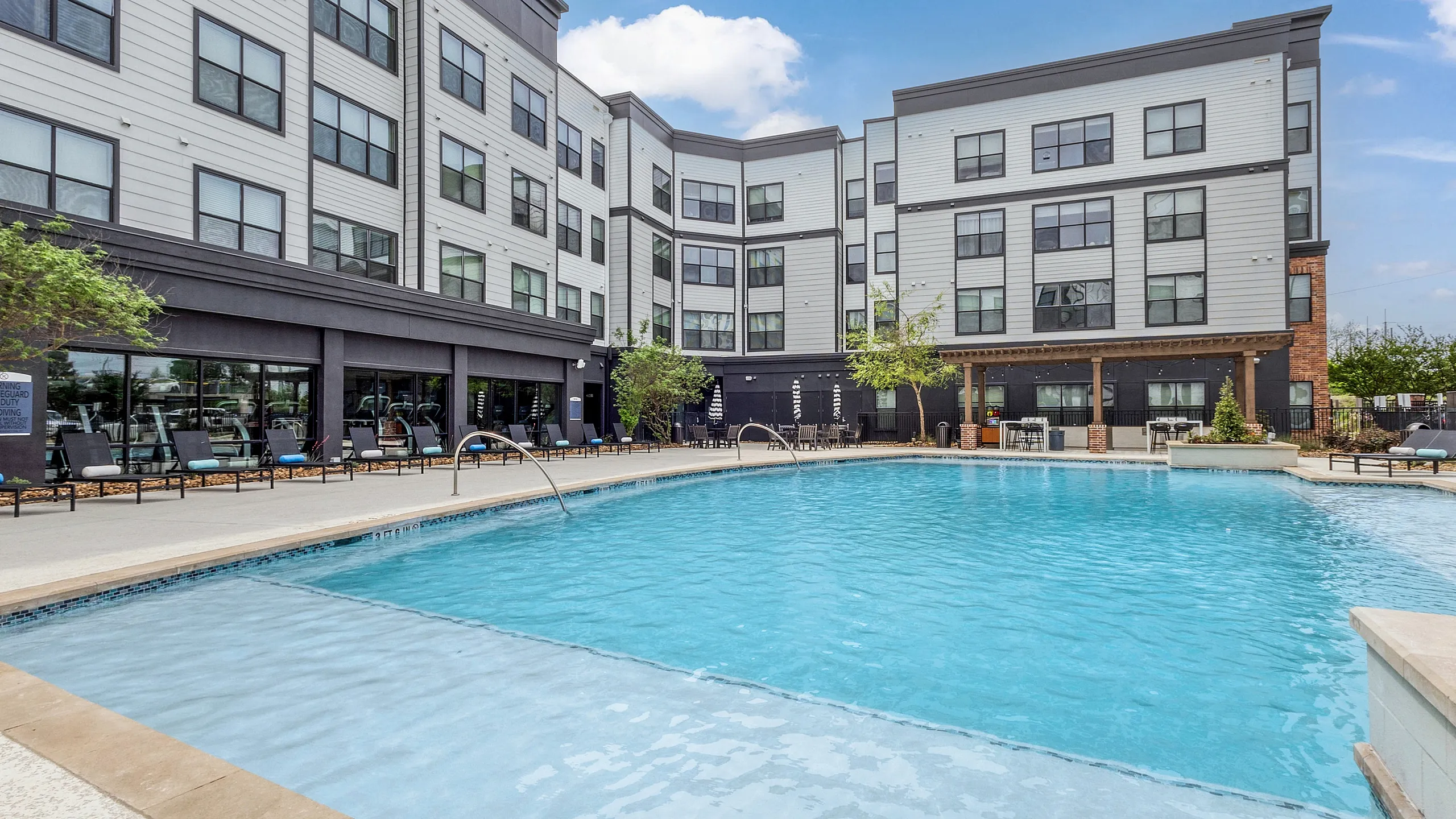 Modern apartment complex featuring a luxurious swimming pool with sun loungers, set against a backdrop of stylish multi-story residential buildings and clear blue skies.