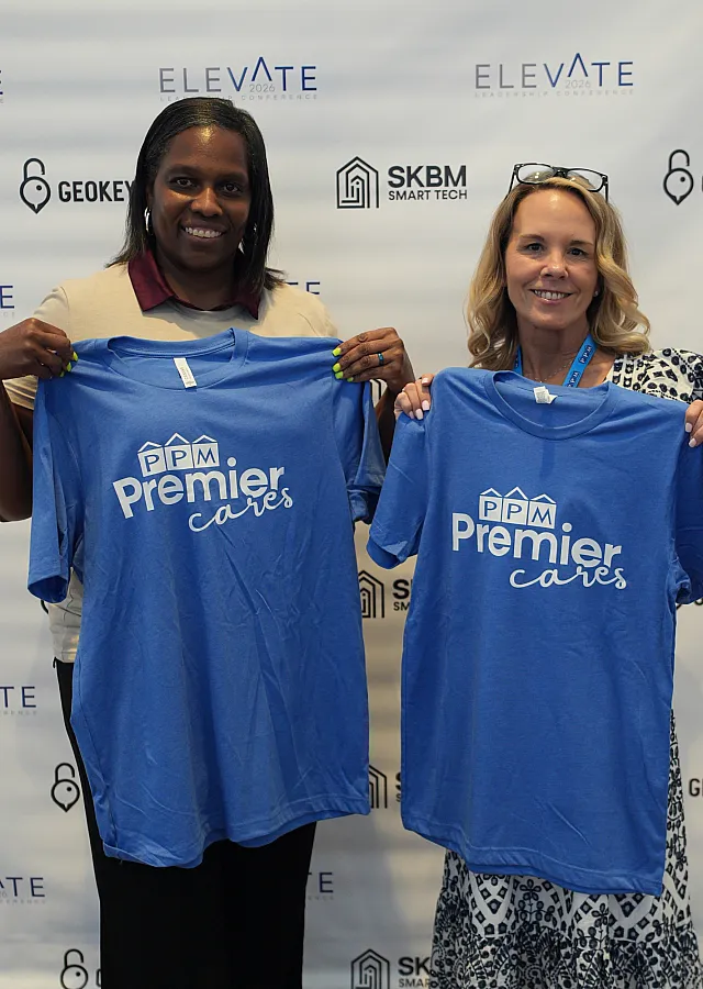 Two smiling individuals holding vibrant blue shirts with 'Premier Cares' logo, standing against a branded wall at an event.